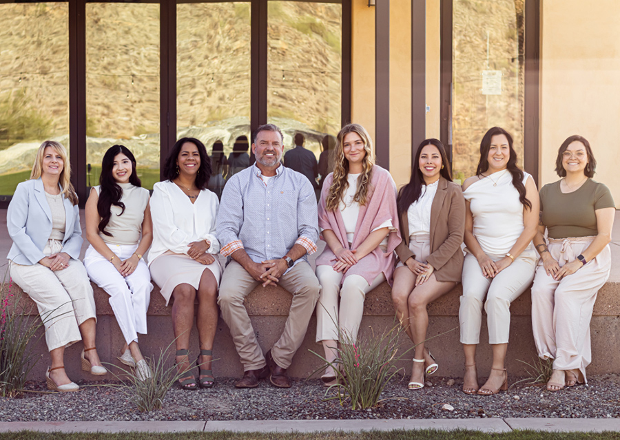 Dental team members seated together in casual attire against a wooden wall backdrop.